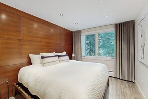 Ein modernes Schlafzimmer mit einem großen Bett, Holzvertäfelung an den Wänden, einem Fenster mit Vorhängen und Blick auf Bäume.