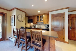 A kitchen with wooden cabinets, a clock, and a bar area with chairs. - Ski-in/out, mountain view condo w/ fireplace, fast WiFi, shared hot tubs/pool/WD (Vail)
