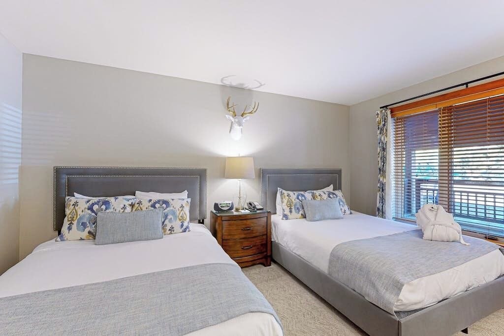 A hotel room with two beds, a nightstand, a lamp, a mounted deer head, and a window with blinds.