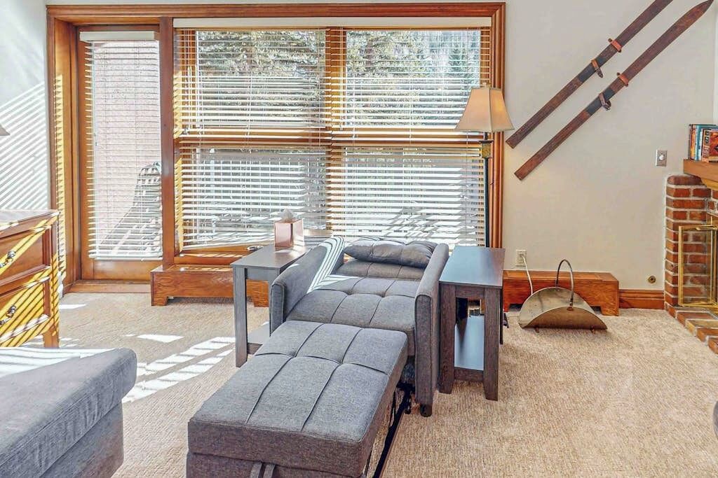 A room with a sofa, ottoman, and a table with a lamp. There is a window with blinds and a staircase in the background.