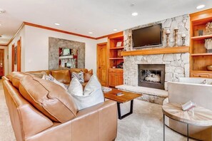 A living room with a stone fireplace, leather sofa, and a flat-screen TV mounted above it. - Ski-in/out condo w/ mountain view, fireplace, fast WiFi, shared pool/hot tubs/WD (Vail)