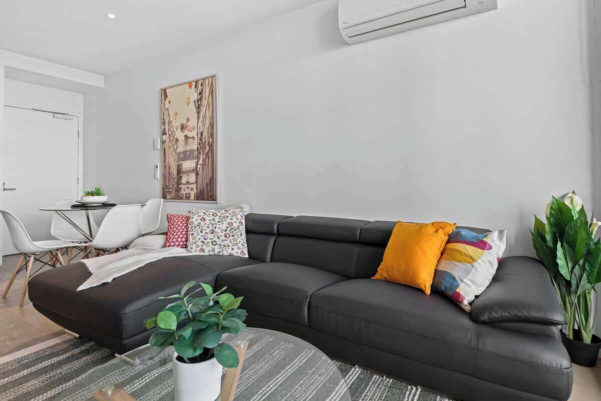Un salon moderne avec un canapé d’angle noir, un coin repas avec des chaises blanches, un tableau au mur et une plante en pot.