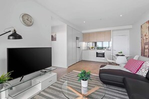 Ein modernes Wohnzimmer mit Flachbildfernseher, Glastisch, Sofa und einem Essbereich mit Küche im Hintergrund.