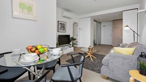 A modern living room with a glass dining table, grey sofa, and a TV. - Caleb - Unique Melbourne Experience *WiFi Park Gym (Melbourne)