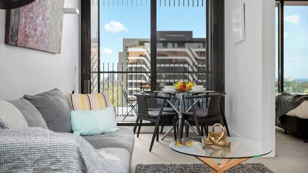 A modern living room with a sofa, a glass coffee table, and a balcony with a view of buildings. - Caleb - Unique Melbourne Experience *WiFi Park Gym (Melbourne)