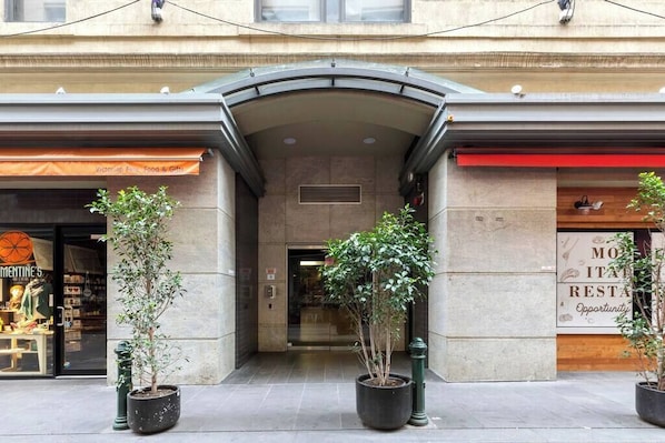 A building entrance with a covered walkway, potted plants, and storefronts including a restaurant and a clothing store. - Xavier - Best location in Melbourne with Parking! (Melbourne)