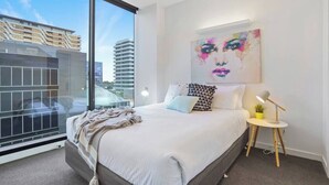 Un dormitorio moderno con una cama grande, una mesita de noche con lámpara, una planta pequeña y vista a un paisaje urbano a través de un ventanal amplio.