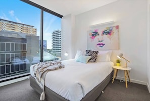 Een moderne slaapkamer met een groot bed, een nachtkastje met een lamp, een kleine plant en een stadsgezicht door een groot raam.