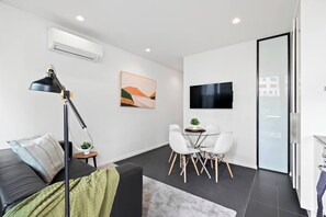 A modern living room with a sofa, a dining table with chairs, a TV, and an air conditioning unit. - Dee - Gorgeous modern apartment in perfect location (South Melbourne)