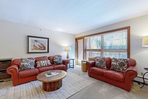 A living room with two red leather sofas, a wooden coffee table, a framed picture on the wall, and a window with blinds. - Ski-in/out, mountain view condo with fireplace & shared pool, hot tub, laundry (Vail)