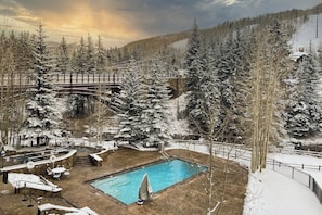 Un área de piscina al aire libre cubierta de nieve con un puente de madera, rodeada de árboles nevados y un paisaje montañoso.