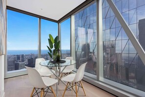Ein moderner Essbereich mit einem Glastisch und weißen Stühlen, großen Fenstern mit Stadtblick und einer Zimmerpflanze.