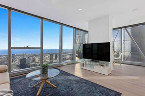 Ein modernes Wohnzimmer mit großen Fenstern, einem Flachbildfernseher, einem Glastisch und Blick auf die Stadt.