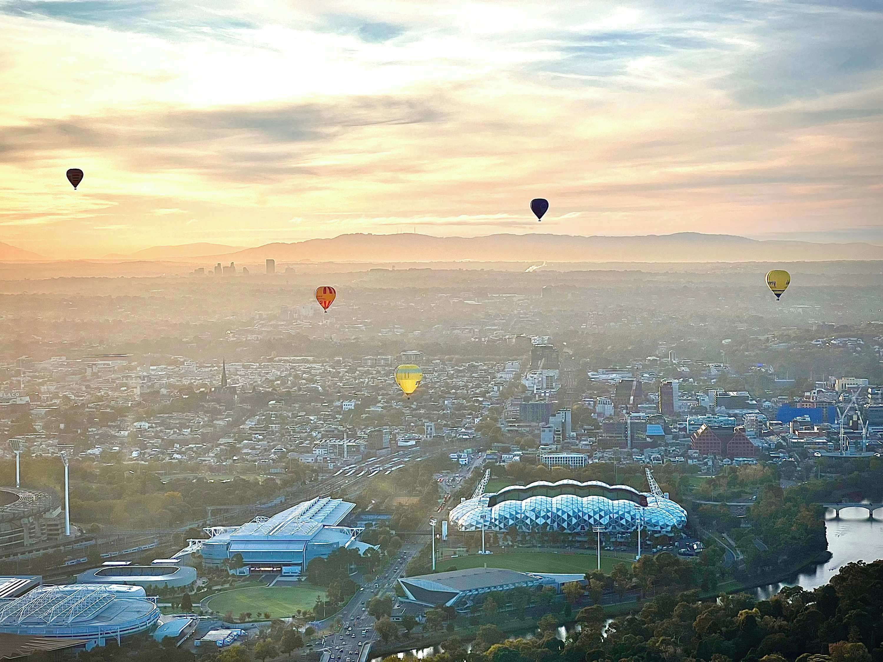 A cityscape with multiple hot air balloons, a stadium, and surrounding buildings.