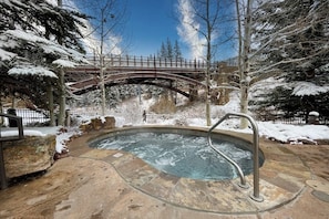 Ein Whirlpool mit Metallgeländer, umgeben von schneebedeckten Bäumen und einer Steinterrasse.