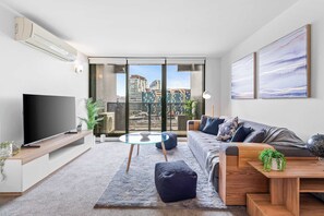 Ein modernes Wohnzimmer mit Flachbildfernseher, Glastisch, Sofa und Blick auf eine Skyline durch große Fenster.