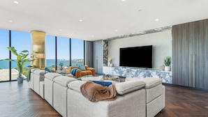 A modern living room with a large sectional sofa, a flat-screen TV mounted on the wall, and a view of the sea through large windows.