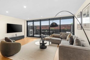 A modern living room with a sofa, armchair, and coffee table, large windows, and a flat-screen TV. - Ultralux City Townhouse with Parking, Gym + Views (West Melbourne)