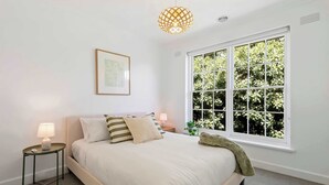 Una camera da letto con un letto, comodini, una lampada a sospensione, una finestra con vista su piante e un quadro appeso al muro.