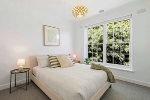 A bedroom with a bed, bedside tables, a hanging light fixture, a window with a view of greenery, and a framed picture on the wall. - Gorgeous Botanic Gardens Retreat with Parking (South Yarra)
