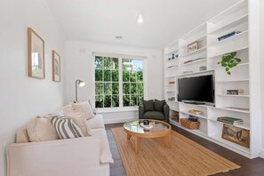 A modern living room with a sofa, a coffee table, a TV, and a bookshelf. - Gorgeous Botanic Gardens Retreat with Parking (South Yarra)