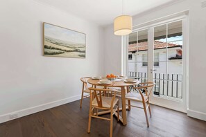 A dining area with a round table, wooden chairs, a hanging light fixture, a painting on the wall, and a view through a glass door. - Gorgeous Botanic Gardens Retreat with Parking (South Yarra)