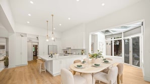 A modern kitchen with white cabinetry, a central island, and a dining area with a round table and chairs. - Luxury Seaside Estate with Pool & Garden Oasis (Brighton)