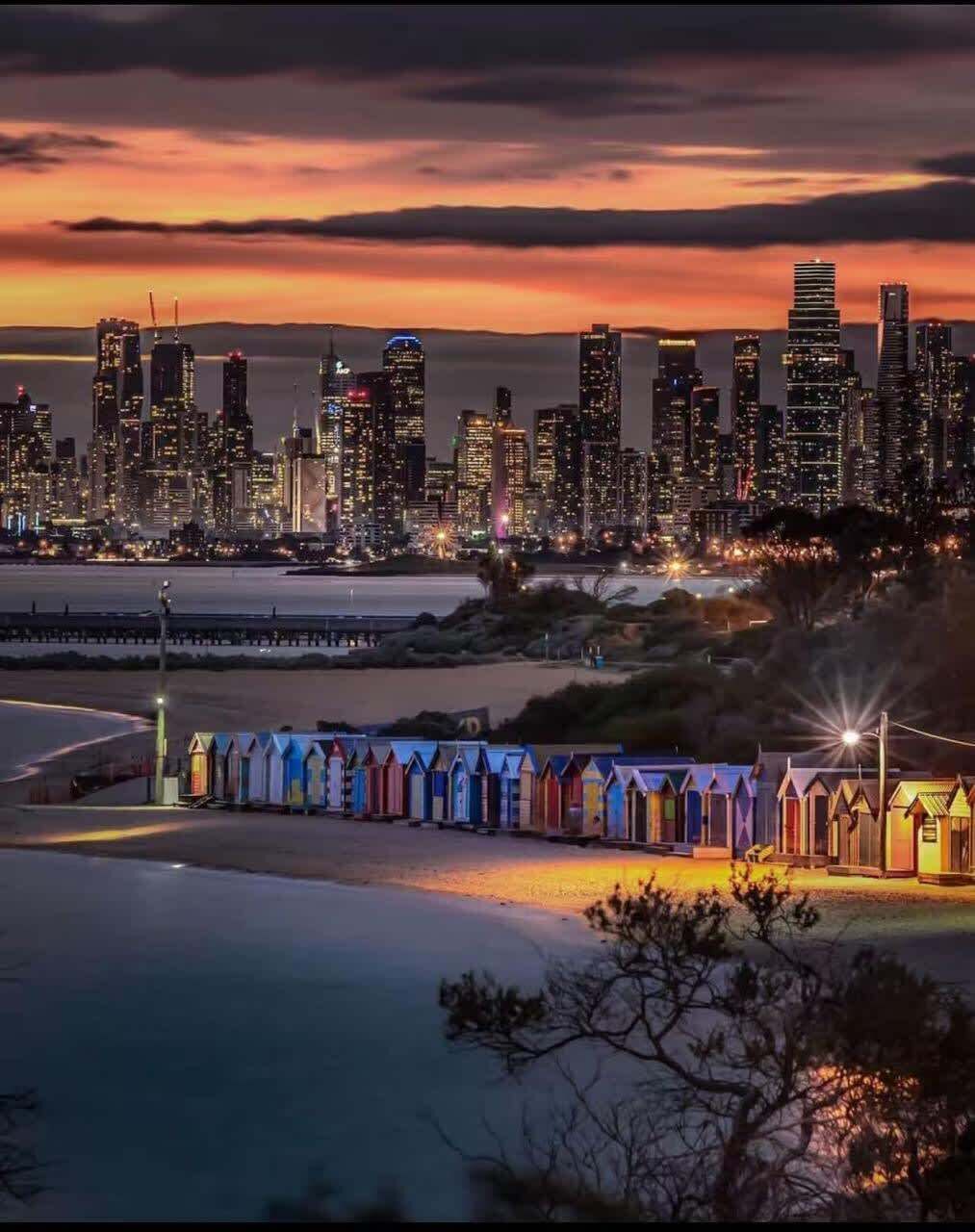 Une vue sur les toits d’une ville au crépuscule, avec des bâtiments illuminés, une plage et une rangée de cabanes de plage aux couleurs vives.