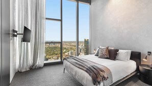 Quarto moderno com uma cama grande, televisão na parede e vista da cidade pelas amplas janelas.