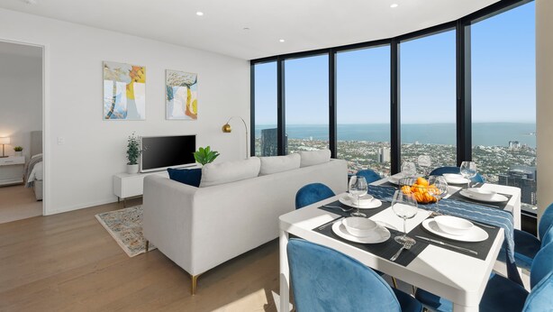 A modern living room with a white sofa, a flat-screen TV, and artwork on the wall. There's a dining area with a table set for two, and a large window offering a city view. - Live in the clouds! 3BD luxury apt. in Southbank. (Southbank)