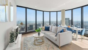 A modern living room with a large window, a white sofa, a dining table set, and a potted plant. - Live in the clouds! 3BD luxury apt. in Southbank. (Southbank)