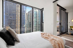 Une chambre avec une grande fenêtre, un lit, une porte coulissante et une vue sur des immeubles de grande hauteur.