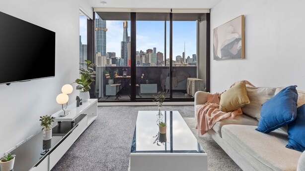 Una sala moderna con televisor grande, sofá, mesita de centro y vista a la ciudad a través de amplias ventanas.