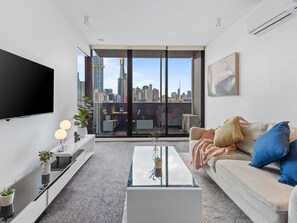 Ein modernes Wohnzimmer mit großem Fernseher, einer Couch, einem Couchtisch und Blick auf die Stadt durch große Fenster.
