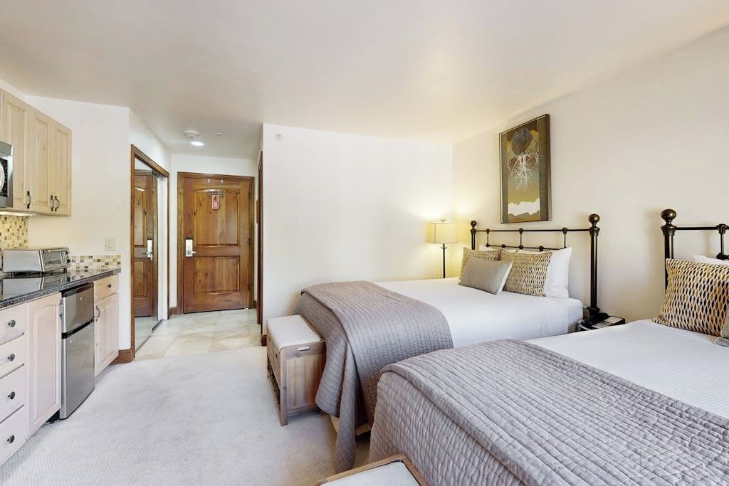 A hotel room with a kitchenette, two beds, a painting on the wall, and a door leading to another room.