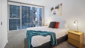 Een slaapkamer met een groot raam, een bed, een nachtkastje en een wanddecoratie.