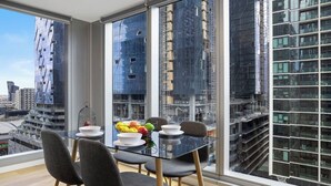 Een moderne eetruimte met een glazen tafel en stoelen, een fruitschaal en uitzicht op een stadsbeeld met hoge gebouwen.