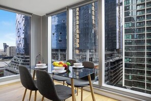 Ein moderner Essbereich mit einem Glastisch und Stühlen, einer Obstschale und Blick auf eine Skyline mit hohen Gebäuden.