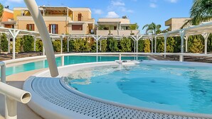 Une piscine dotée d’une terrasse blanche, entourée d’immeubles avec des balcons, sous un ciel dégagé.