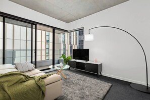 Ein modernes Wohnzimmer mit einer Couch, einem Glastisch, einem Fernseher auf einem schwarzen Schrank und einem großen Fenster.