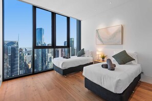 Ein modernes Hotelzimmer mit zwei Betten, einem großen Fenster mit Blick auf die Stadt, einem Wandbild und einem kleinen Nachttisch mit Lampe und Pflanze.