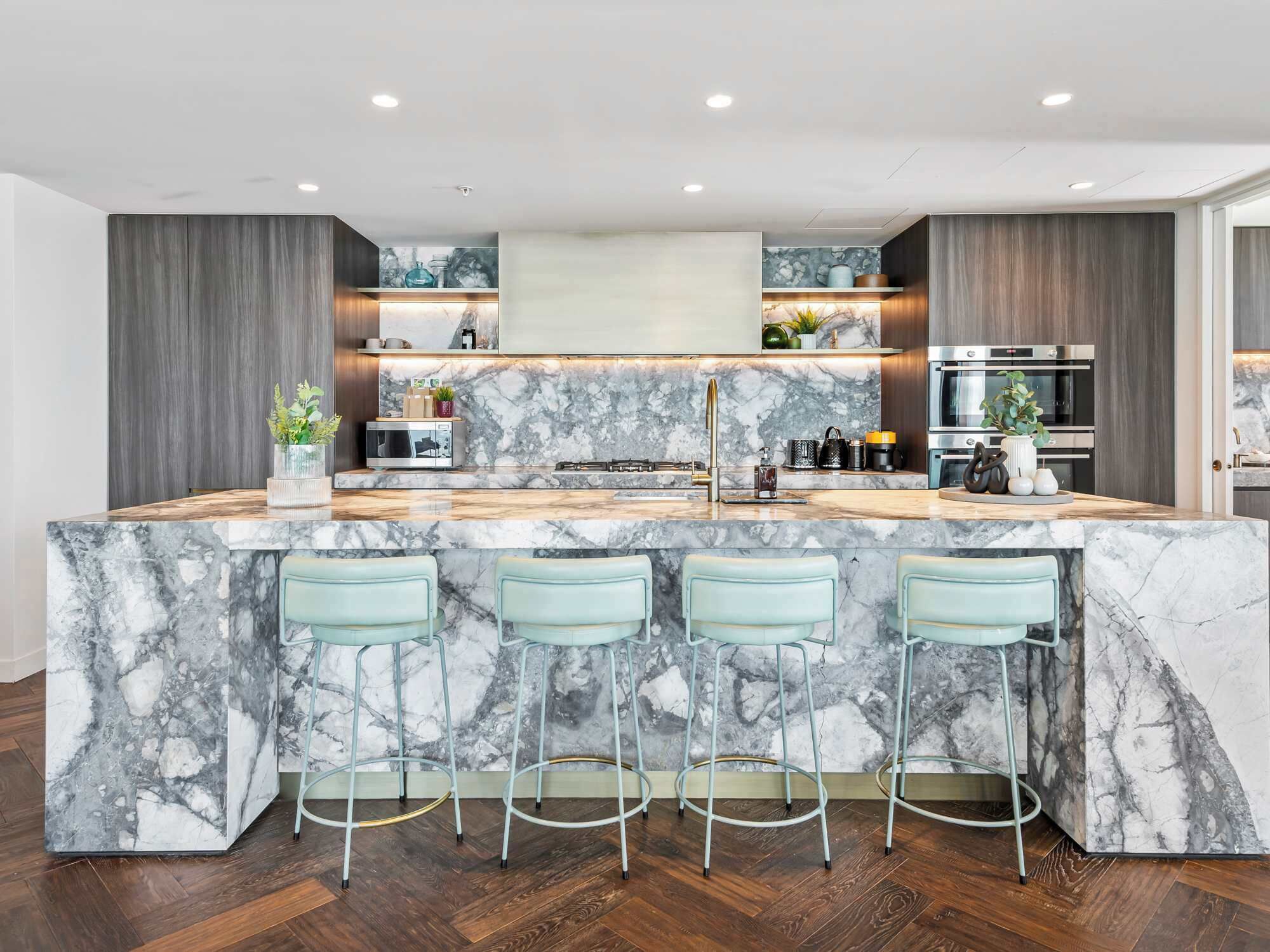 A modern kitchen with a marble island, bar stools, built-in appliances, and a herringbone wood floor.