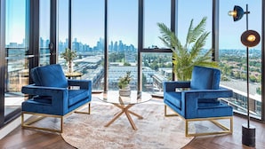 Una stanza angolare con due poltrone blu, un tavolino rotondo in vetro, un'ampia finestra con vista sulla città e una lampada da terra.