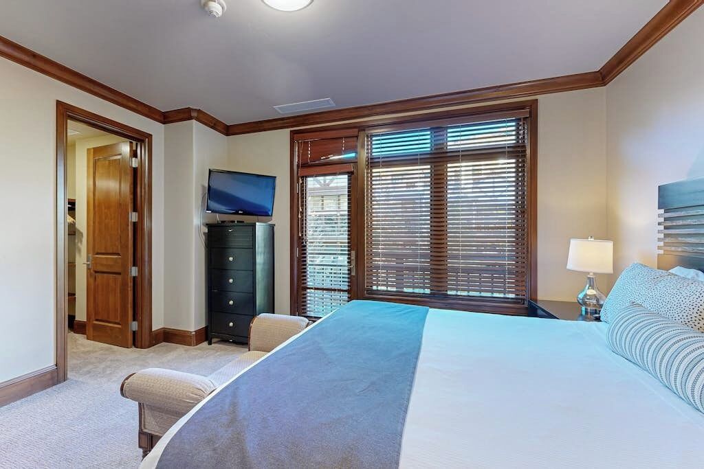 A bedroom with a bed, a TV, a dresser, and a window with blinds.