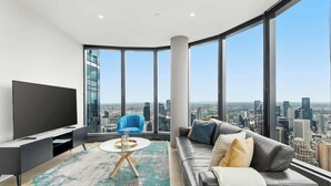 Un moderno salón con televisor grande, una butaca azul, un sofá gris y vista a la ciudad a través de amplios ventanales.