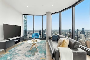 Ein modernes Wohnzimmer mit großem Fernseher, einem blauen Sessel, einem grauen Sofa und Blick auf die Stadt durch große Fenster.