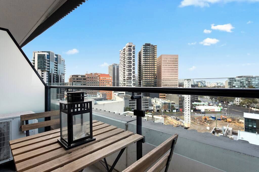 A balcony with a wooden table and a black lantern, overlooking a cityscape with various buildings.