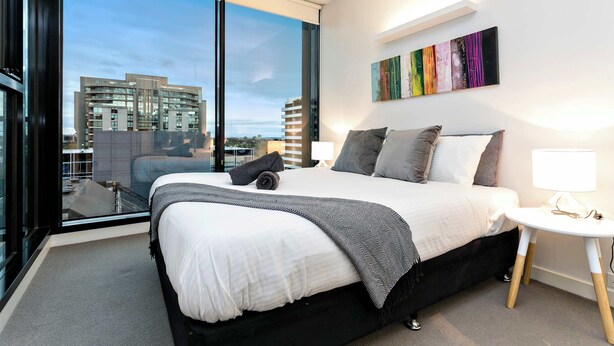 A modern hotel room with a large bed, a bedside table, a view of the city, and a colorful abstract painting on the wall. - Nora - Superb designer style *WIFI Parking (Melbourne)