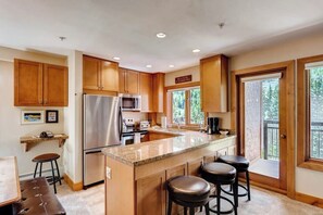 A kitchen with wooden cabinets, a stainless steel refrigerator, a granite countertop, and a window overlooking greenery. - Ski-in/out Condo w/ Three Decks, Fireplace, WiFi & Shared Pool/Hot Tub/Laundry (Vail)