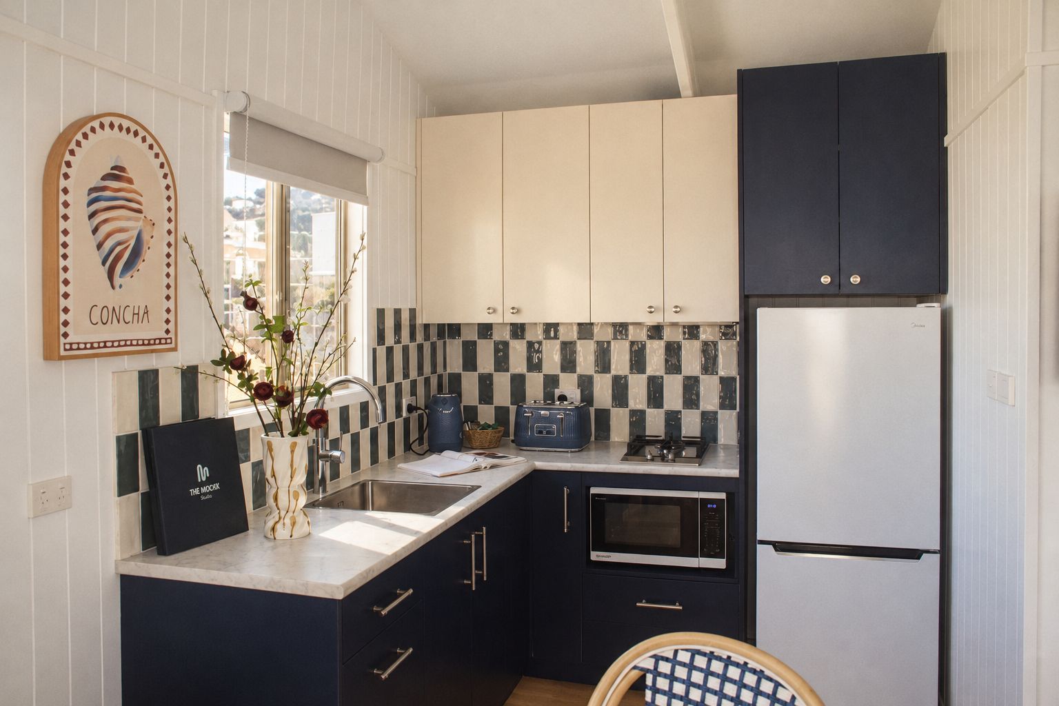 A compact kitchen with a sink, stove, and refrigerator, featuring a checkered backsplash and dark cabinetry.